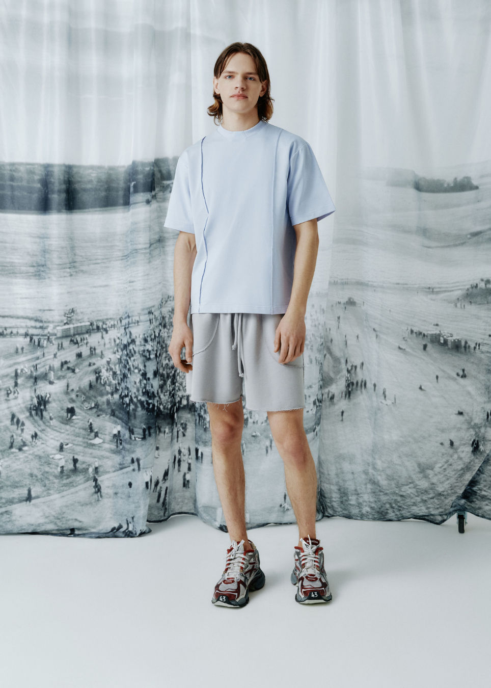 A model stands in a studio wearing a light blue short-sleeve T-shirt, light gray shorts, and patterned sneakers, posed in front of a large black-and-white scenic backdrop.
