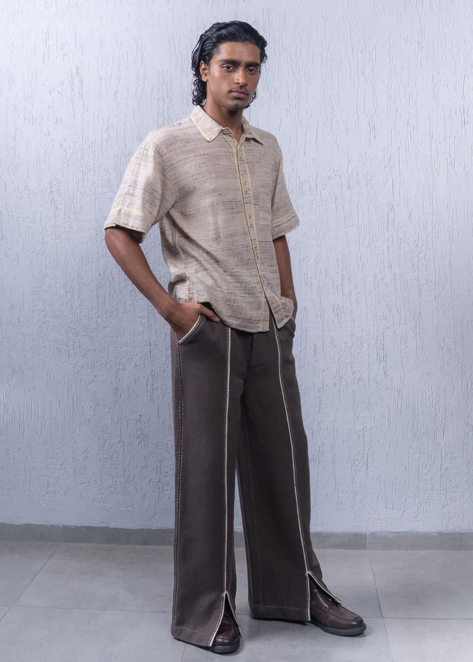 A man modeling a short-sleeve beige textured button-up shirt and dark brown wide-leg trousers with light piping details, standing against a textured light gray wall.