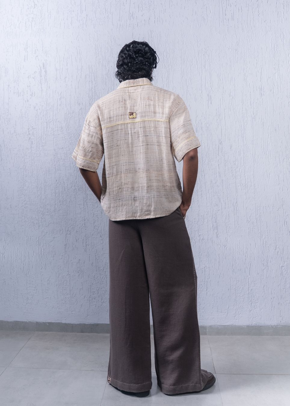 A person standing with their back to the camera, wearing a beige short-sleeve textured button-up shirt and dark brown wide-leg trousers, set against a light gray textured wall.