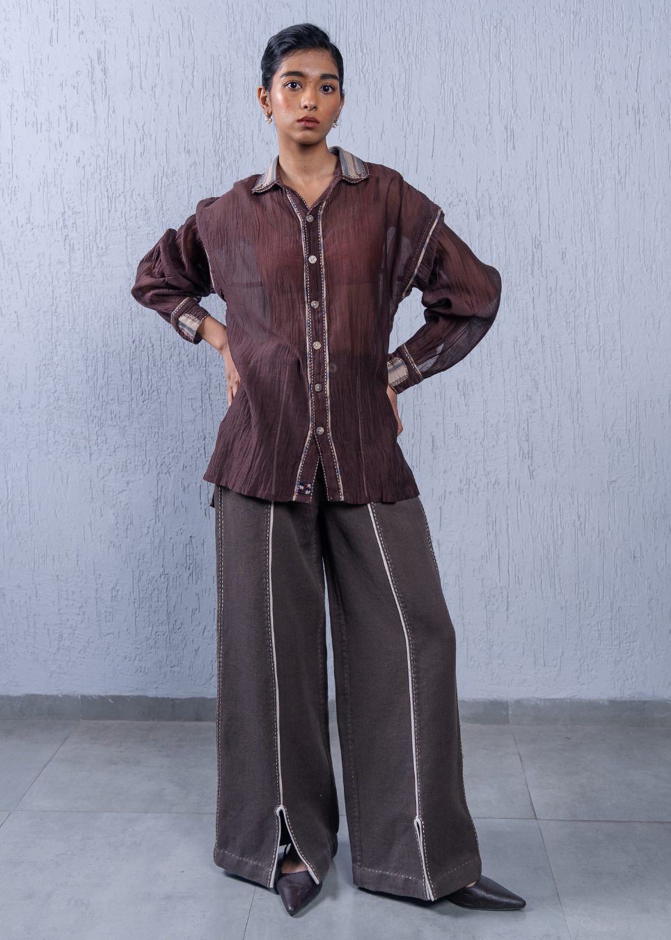 A model stands against a textured light gray wall wearing a dark brown pleated button-up shirt with contrasting light trim and matching wide-leg trousers with vertical light piping.