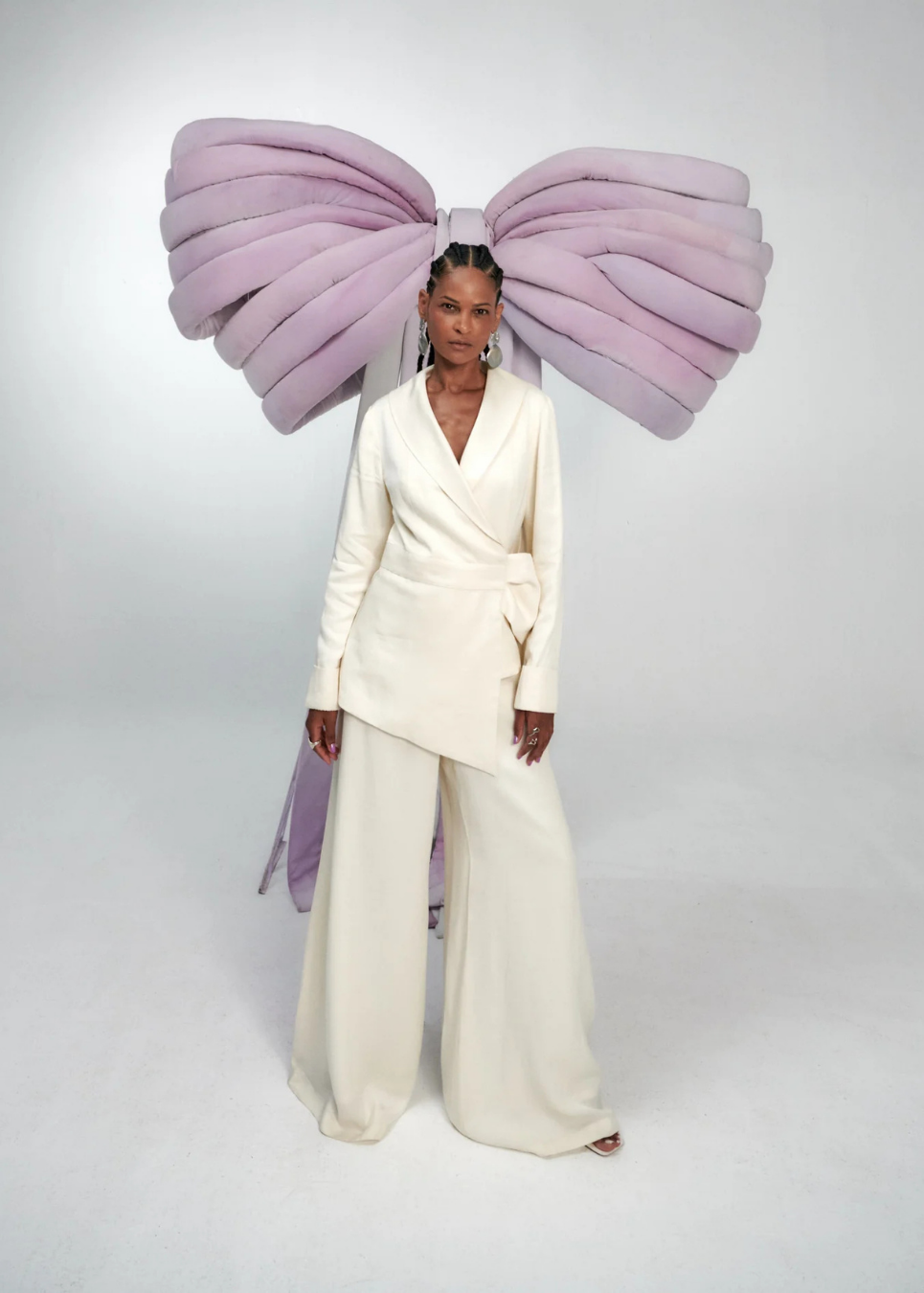 A model stands against a light gray studio background wearing a cream-colored wrap blazer and matching wide-leg trousers, with a dramatic oversized lavender bow headpiece behind her head.