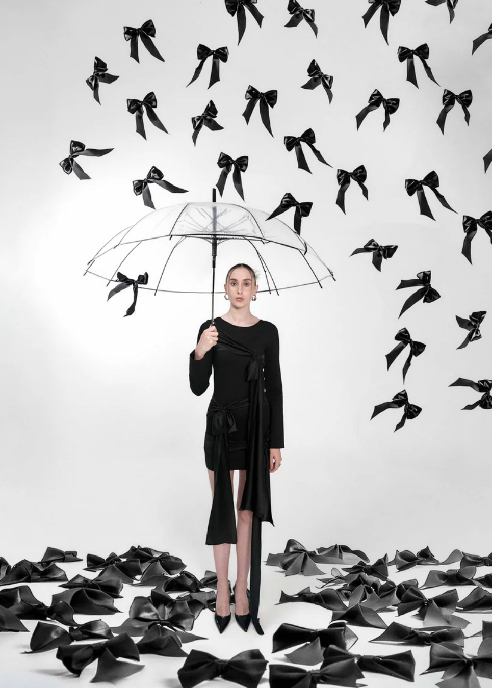 A model in a black dress stands holding a clear umbrella, surrounded by numerous black fabric bows floating in the air and scattered on the floor against a white studio background.