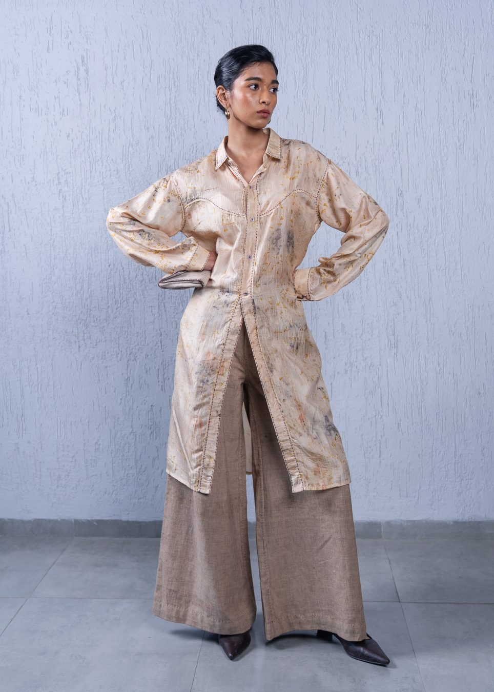 A model stands against a textured light gray wall wearing a long-sleeved, button-front shirt jacket in a beige and brown abstract pattern, paired with wide-leg brown trousers and dark pointed shoes.