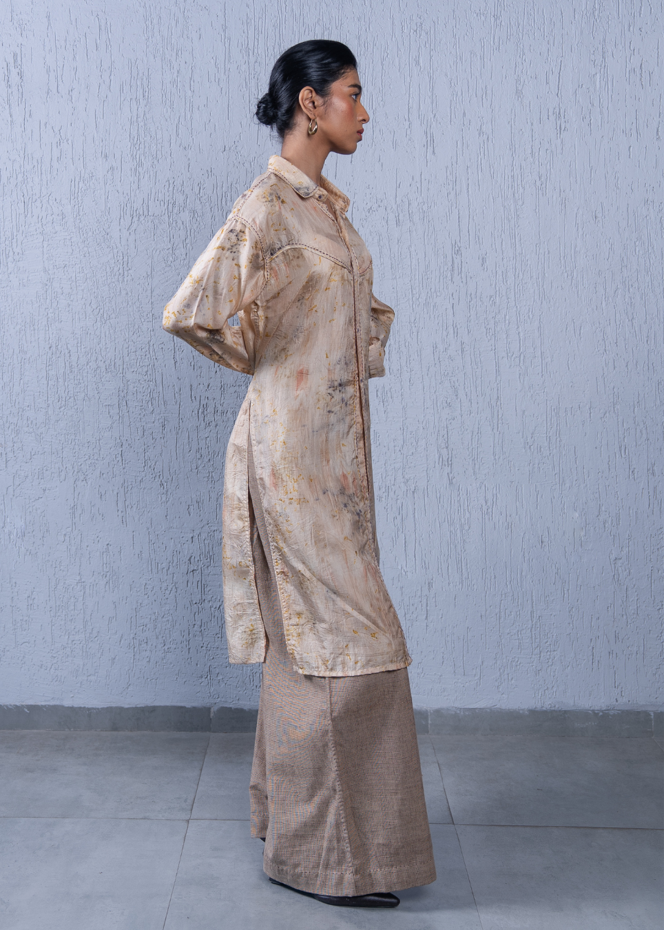 A woman stands in profile wearing a beige and brown floral printed long kurta over a matching wide-leg trouser set, set against a textured light gray wall.