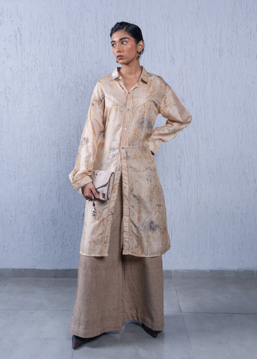 A woman models a long-sleeved beige button-up shirt jacket with a subtle floral pattern, paired with wide-leg brown trousers, standing against a textured light gray wall.