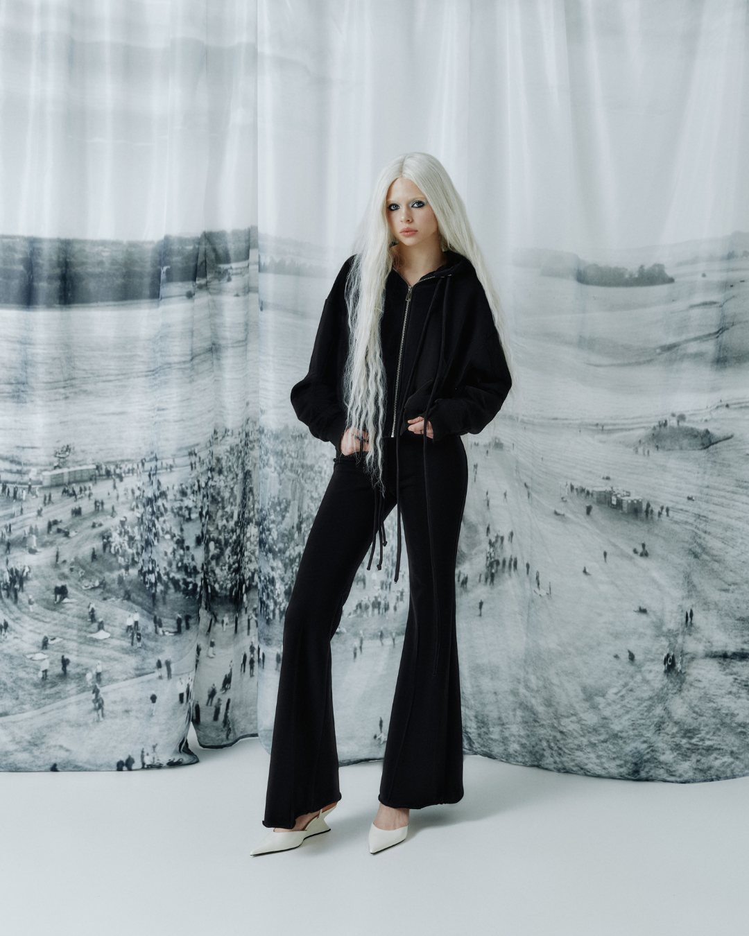 A model with long platinum blonde hair stands in a studio wearing a black zip-up hoodie and matching black flared pants, paired with white pointed shoes. The background is a large draped fabric print featuring a black and white landscape scene.