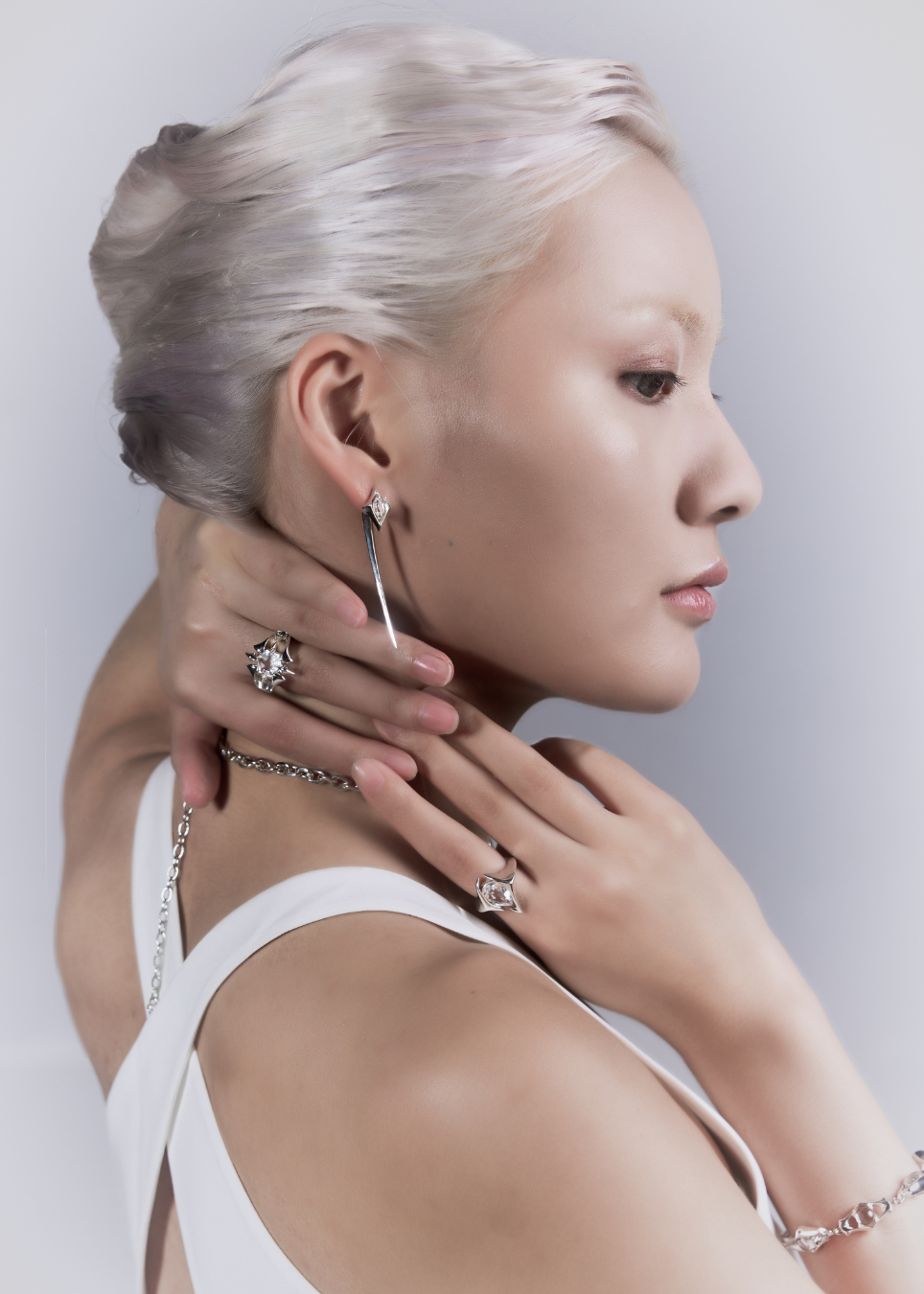 A woman with platinum blonde hair shown in profile wearing delicate silver jewelry, including a long drop earring, rings, and a bracelet, against a light gray studio background.