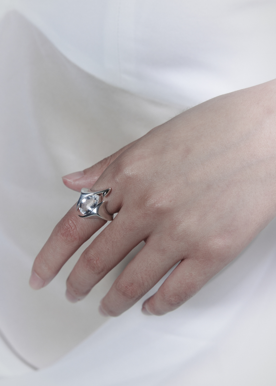 A silver ring with a large, irregularly shaped clear gemstone displayed on a person's finger against a soft white background.