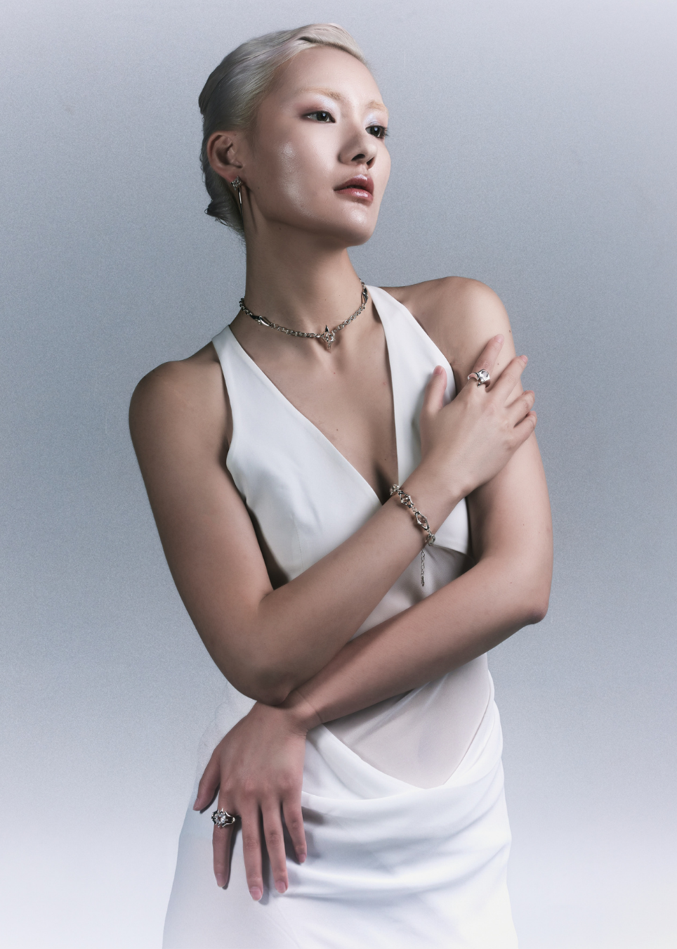 A woman with platinum blonde hair styled in a low bun, wearing a white sleeveless draped dress and silver jewelry, posing against a light gray studio background.