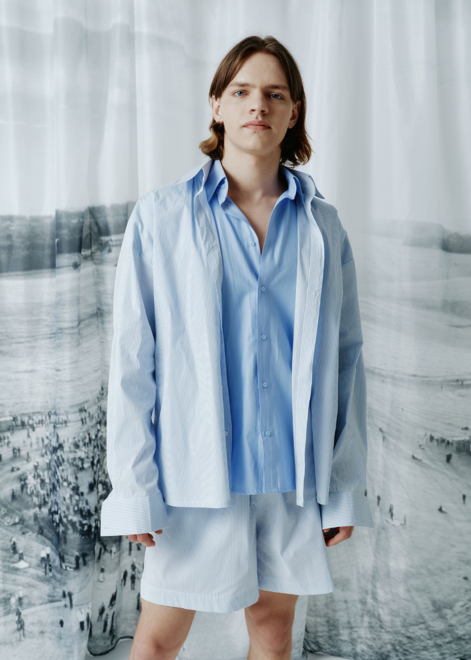 A model stands wearing a light blue button-up shirt layered over a darker blue collared shirt, paired with matching light blue shorts, set against a large black and white scenic backdrop.