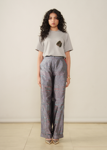 A model stands against a plain beige studio wall wearing a light gray t-shirt with a dark graphic on the chest and high-waisted, wide-leg gray trousers with a subtle pink and blue pattern.