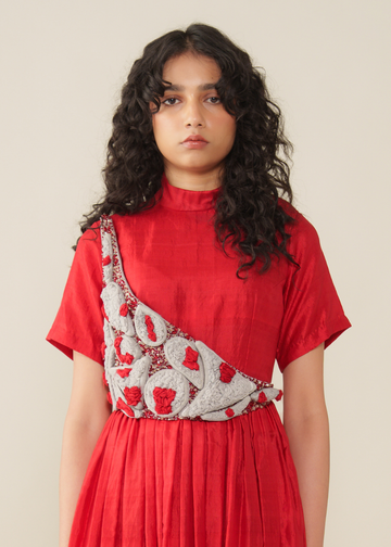 A model with long, curly dark hair wearing a red short-sleeve mock-neck dress with a unique silver and red embroidered diagonal sash detail across the chest, set against a plain beige background.