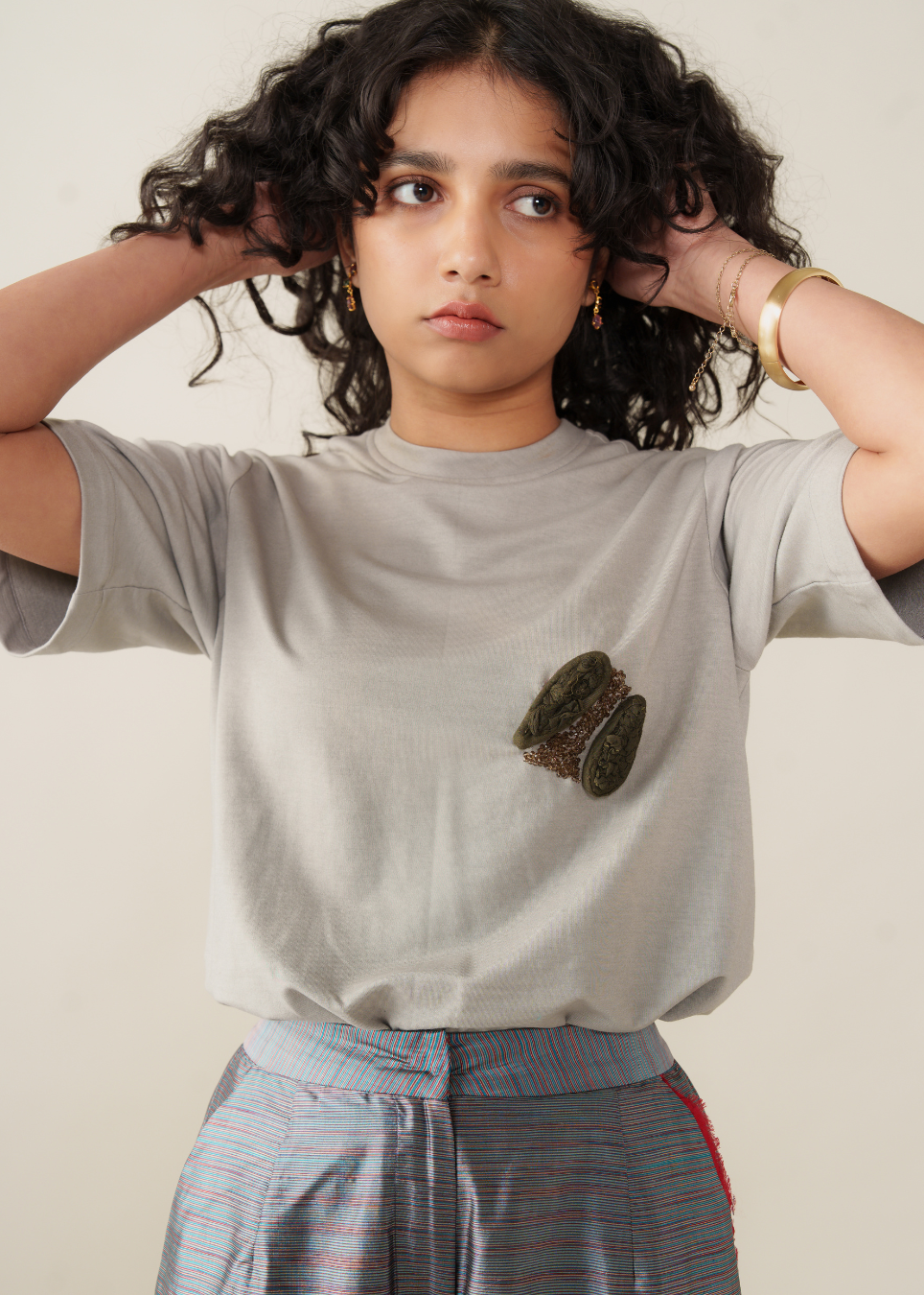 A model with curly dark hair wearing a light gray t-shirt with a small green and brown textured graphic on the left chest, paired with blue and red striped pants, against a plain beige background.