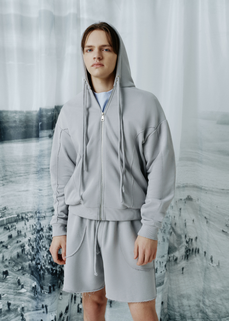 A model wearing a light gray zip-up hoodie and matching drawstring shorts, standing in front of a large, desaturated landscape photograph backdrop.