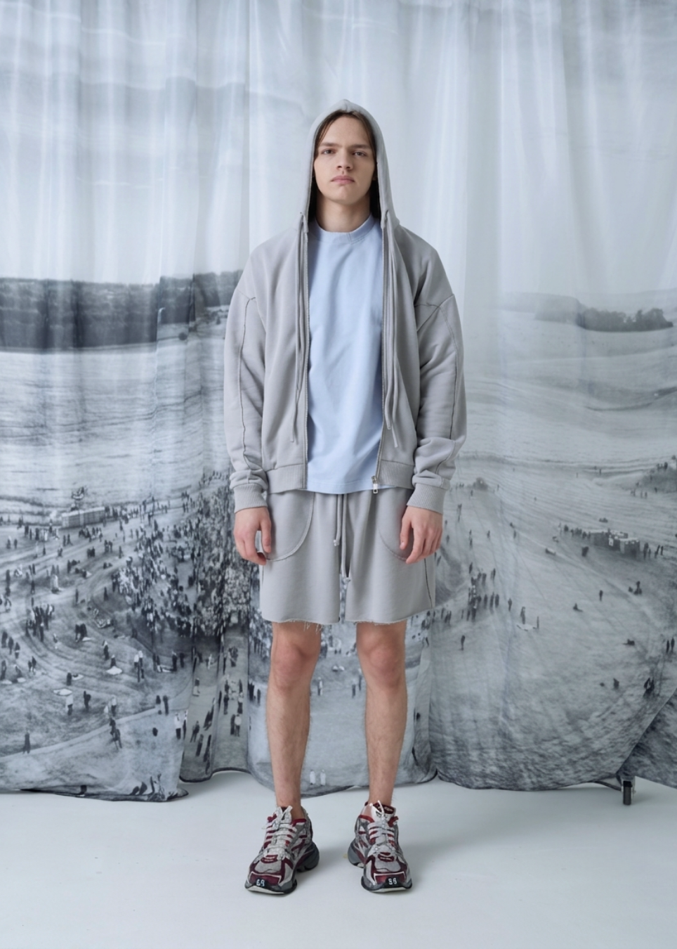 A person stands facing forward wearing a light gray zip-up hoodie, matching gray shorts, a pale blue T-shirt, and patterned sneakers in front of a large black-and-white scenic backdrop.