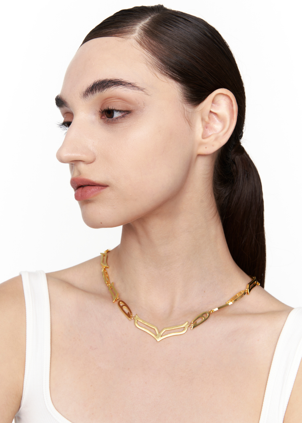 A woman with dark hair in a ponytail wearing a gold chevron necklace with amber-colored oval links, shown in a close-up profile against a white background.