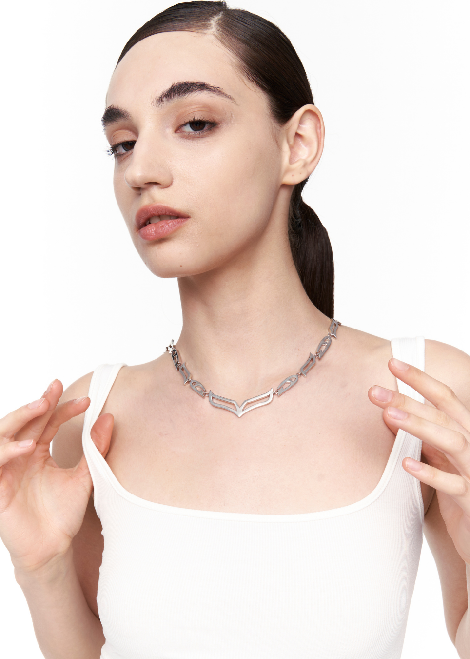 A woman with dark hair tied back wears a silver geometric chain necklace with interlocking chevron links, paired with a white scoop-neck top against a plain white background.
