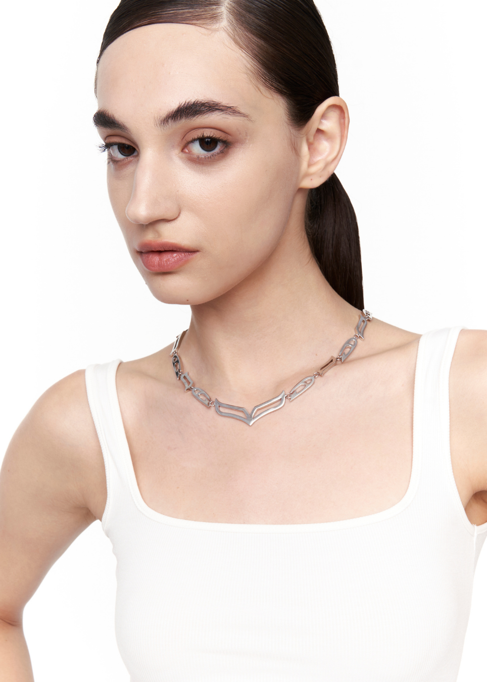 A close-up of a model wearing a silver-toned geometric chain necklace with a textured chevron design, paired with a white tank top against a plain white background.