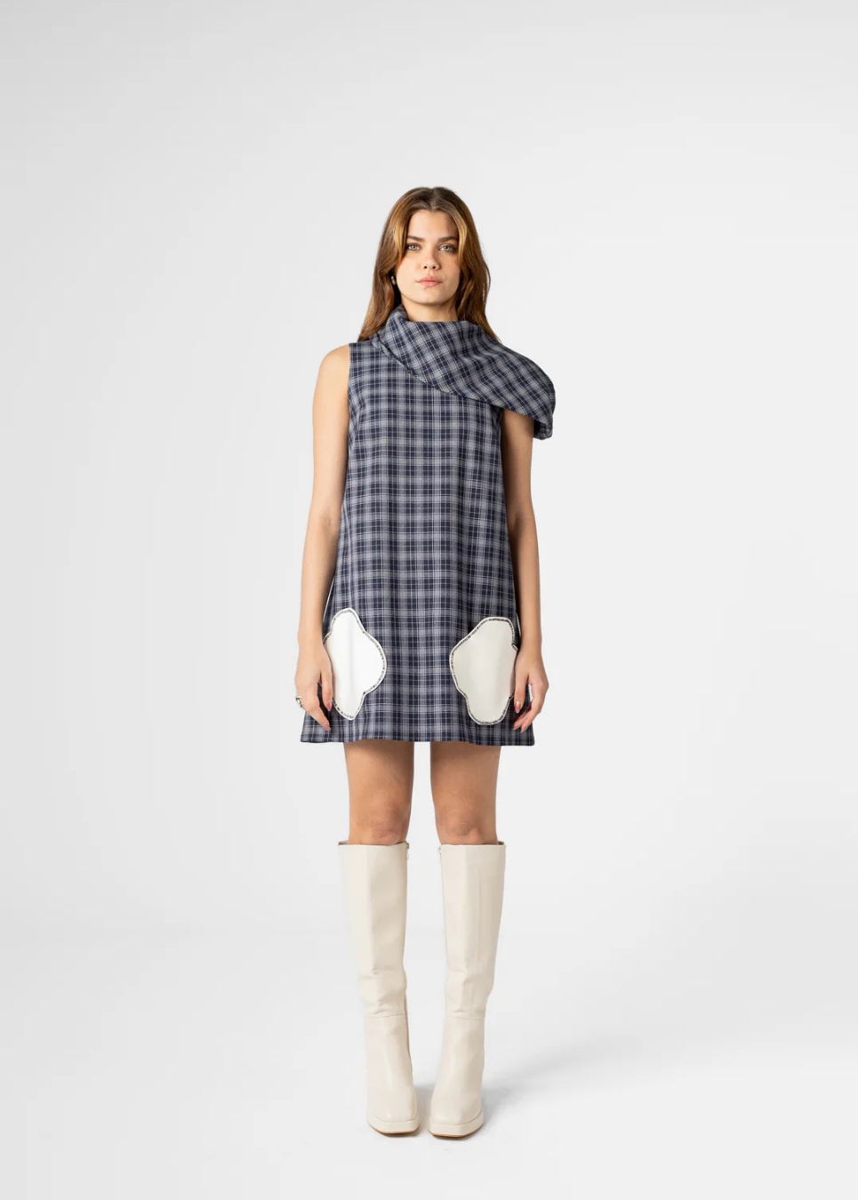 A model wearing a sleeveless plaid mini dress with a unique draped collar and contrasting white elbow patches, paired with cream-colored knee-high boots, against a light gray studio background.