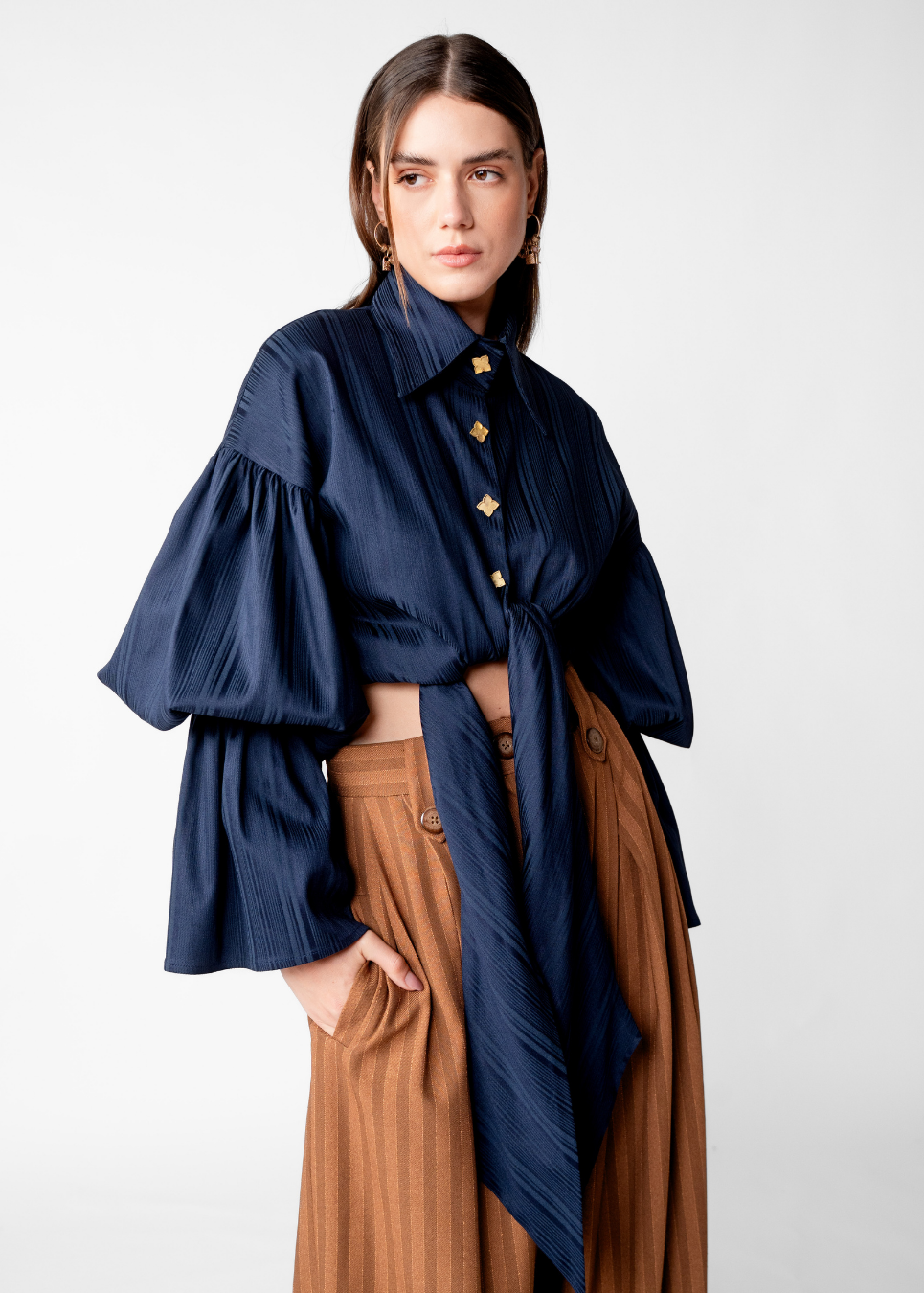 A woman modeling a navy blue long-sleeve blouse with dramatic ruffled bell sleeves and gold-tone buttons, paired with brown pleated trousers, against a light gray studio background.