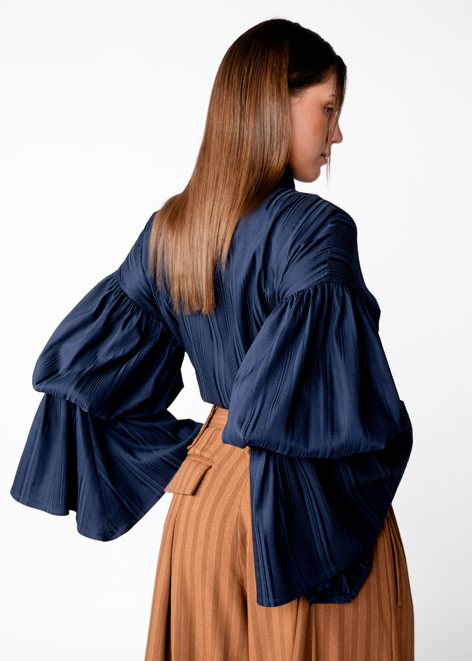 A woman shown from the back wearing a navy blue pleated blouse with dramatic, voluminous bell sleeves and a brown pleated skirt, set against a plain white studio background.