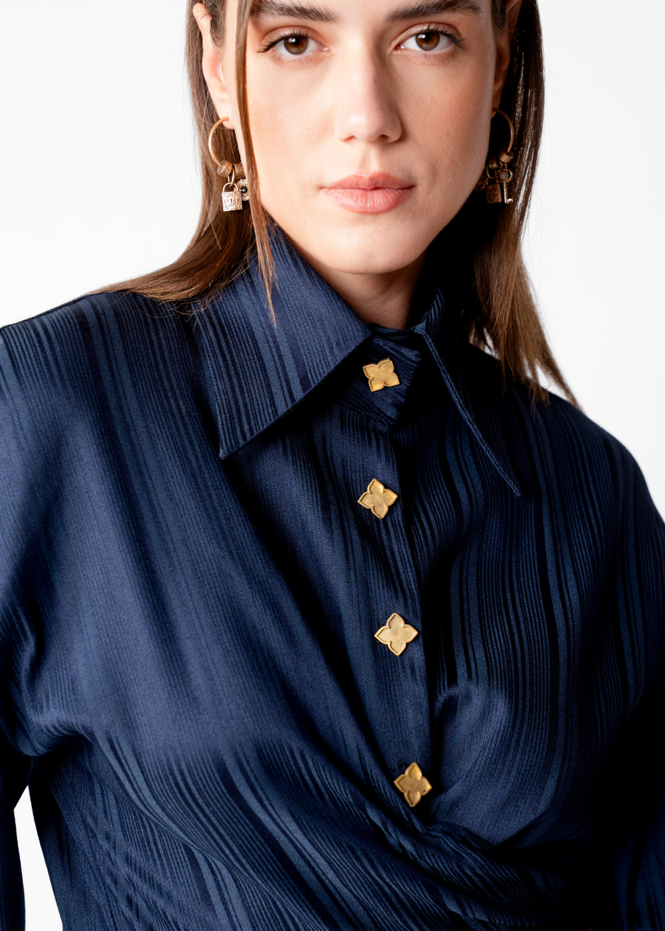 A close-up of a woman wearing a dark navy pleated button-down shirt with decorative gold buttons, set against a plain white background.