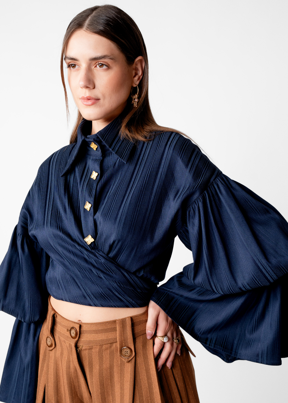 A woman wearing a navy blue pleated blouse with dramatic bell sleeves and gold-tone buttons, paired with brown trousers, against a plain light gray studio background.