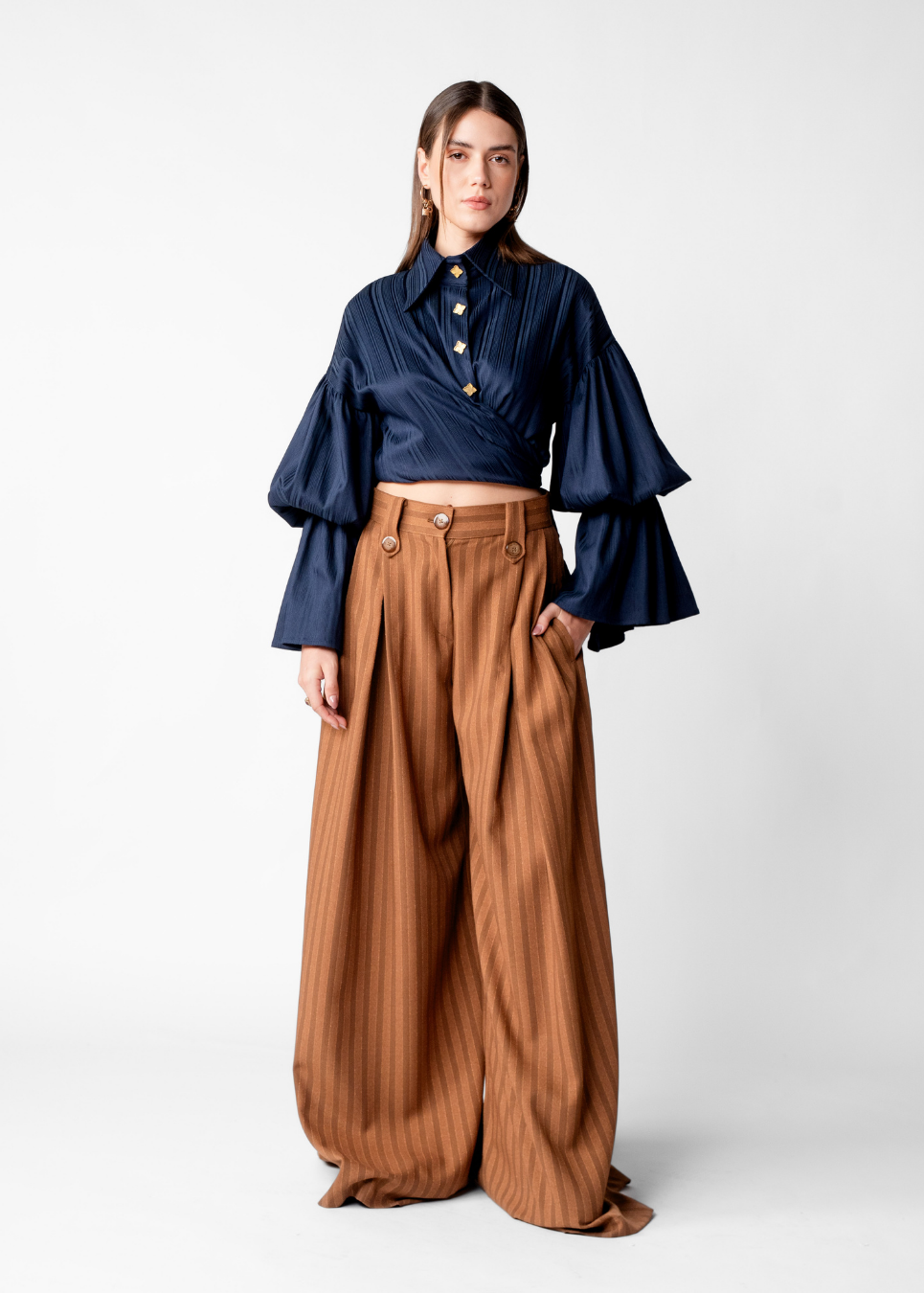 A model stands against a light gray studio background wearing a navy blue long-sleeve blouse with pleated detailing and wide flared bell sleeves, paired with high-waisted, wide-leg brown trousers with vertical pleats and front buttons.