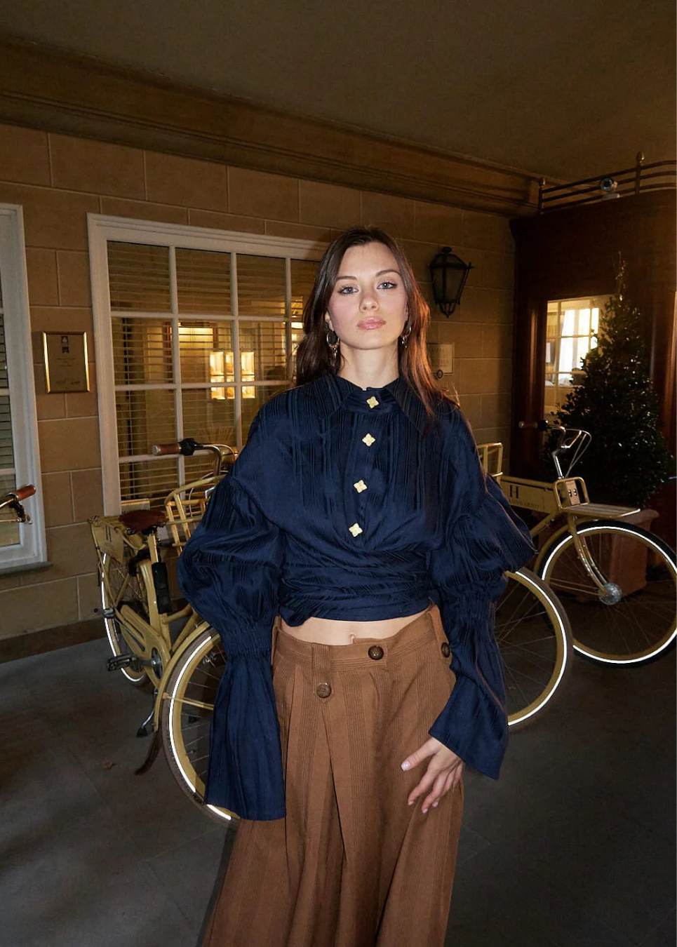 A woman stands indoors wearing a dark navy long-sleeve blouse with gold buttons and brown wide-leg pants. Bicycles and a decorated Christmas tree are visible in the background.