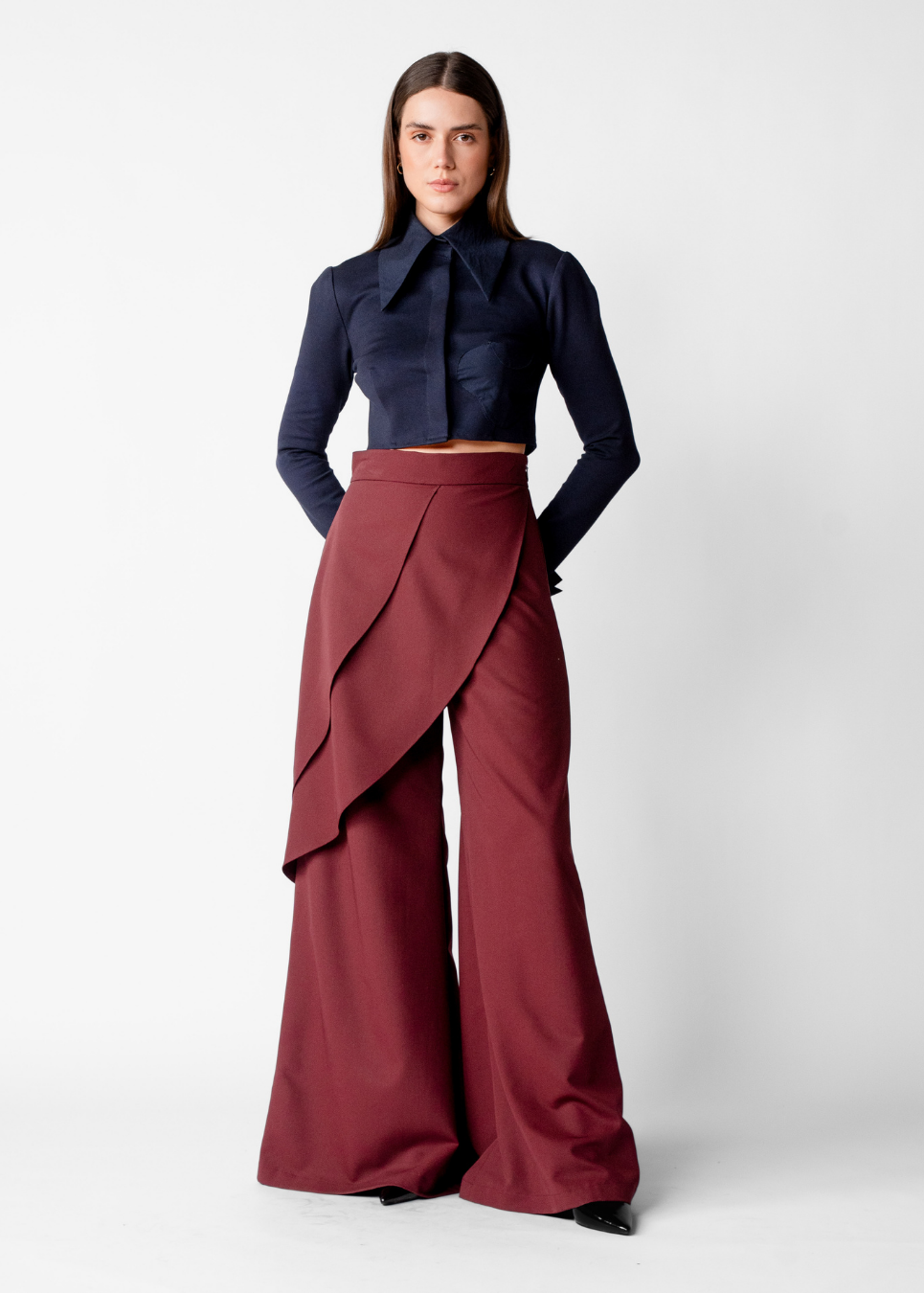 A model stands against a light gray studio background wearing a dark navy cropped long-sleeve blouse with a large bow collar and high-waisted, wide-leg burgundy wrap-style trousers.