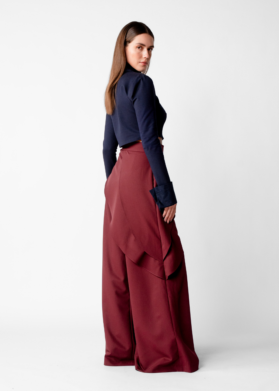 A woman modeling a dark navy long-sleeve top and high-waisted, wide-leg burgundy trousers, photographed from the back against a plain light gray studio background.