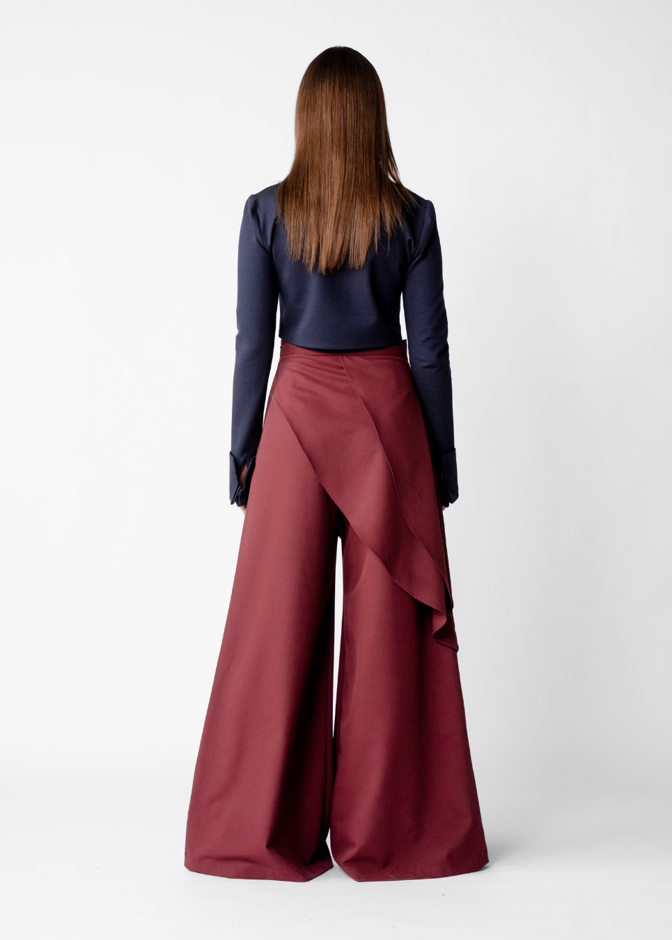 A woman viewed from the back wearing a navy blue long-sleeve top and high-waisted, wide-leg burgundy trousers with an asymmetrical draped detail on the side, set against a plain light gray studio background.