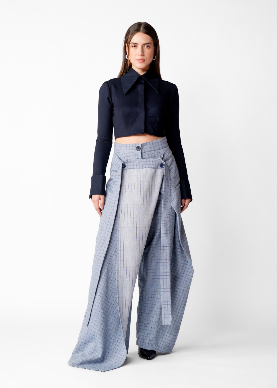 A model stands against a plain white studio background wearing a dark navy long-sleeve cropped collared top and high-waisted wide-leg trousers in a light blue and white check pattern with a draped front detail.