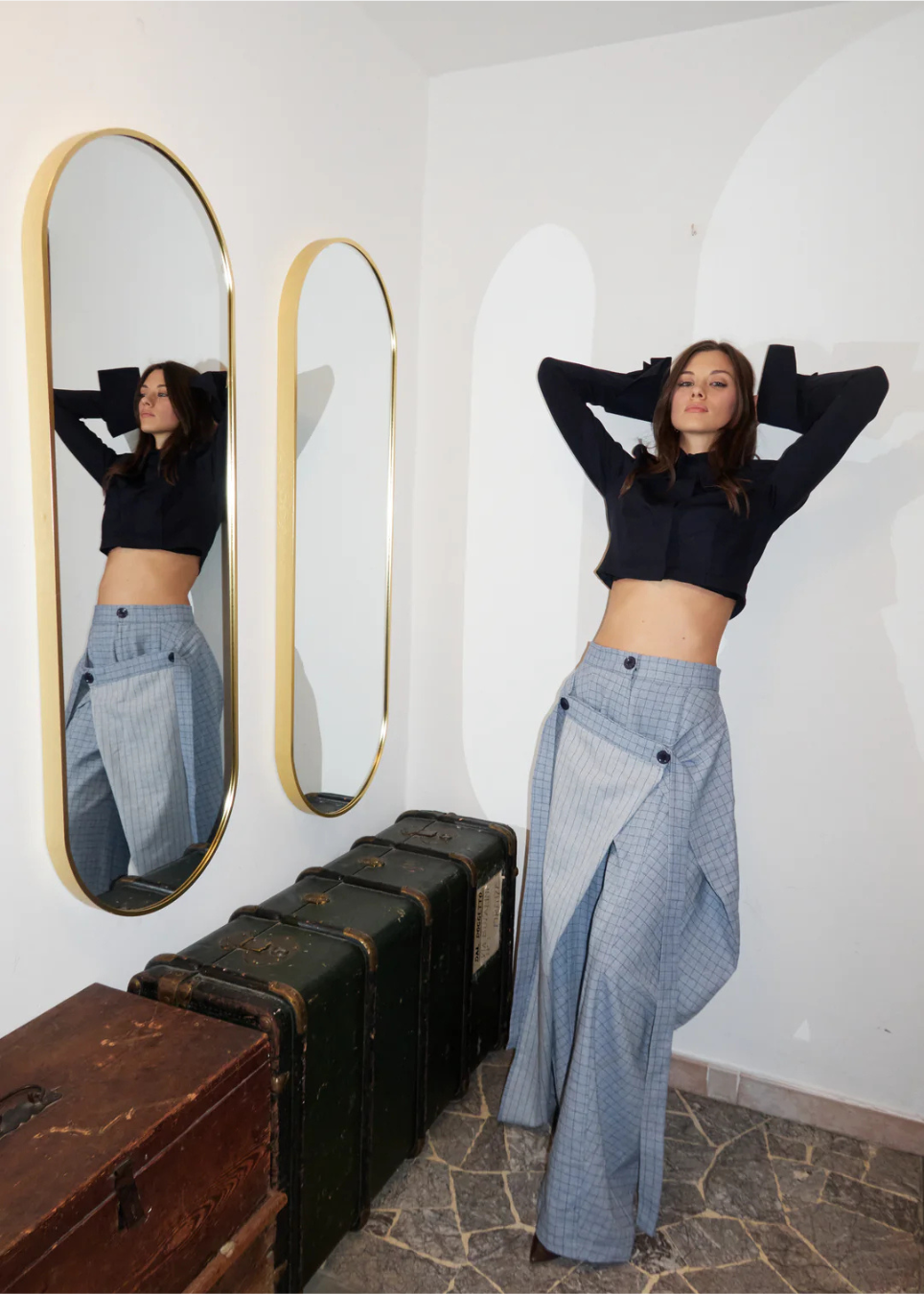 A woman poses in a room wearing a black cropped long-sleeve top and a light blue patterned wrap skirt, with two arched mirrors and vintage suitcases visible.
