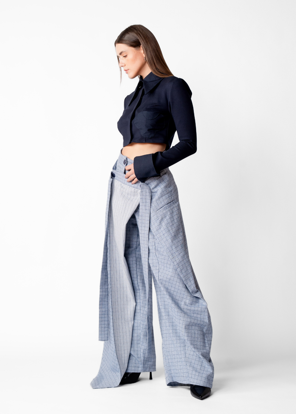 A model is shown from the side wearing a dark navy blue cropped long-sleeve top and wide-leg light blue and white checkered trousers, standing against a plain white studio background.