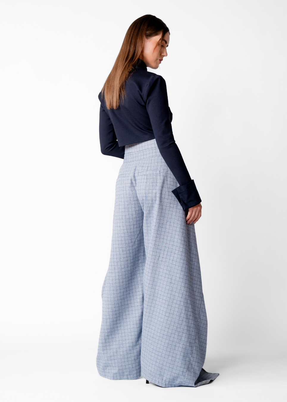 A woman stands in profile wearing a navy long-sleeve top and light blue wide-leg trousers with a subtle grid pattern, photographed against a plain white studio background.