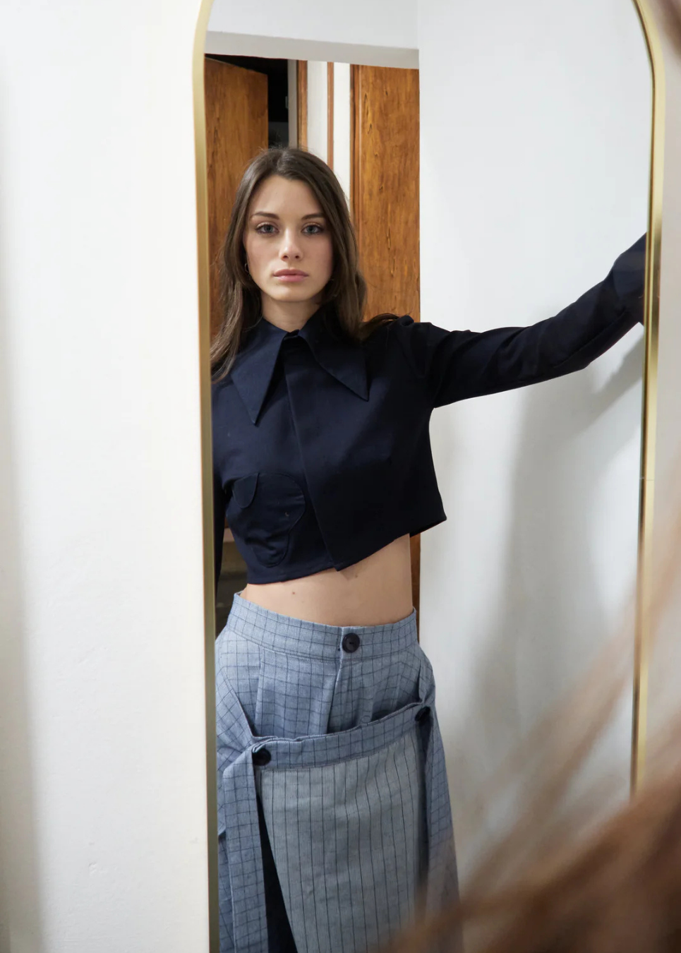 A woman stands in front of a mirror wearing a cropped black long-sleeve collared shirt and a gray pinstriped skirt. The photo is taken indoors with a white wall and wooden door visible in the background.