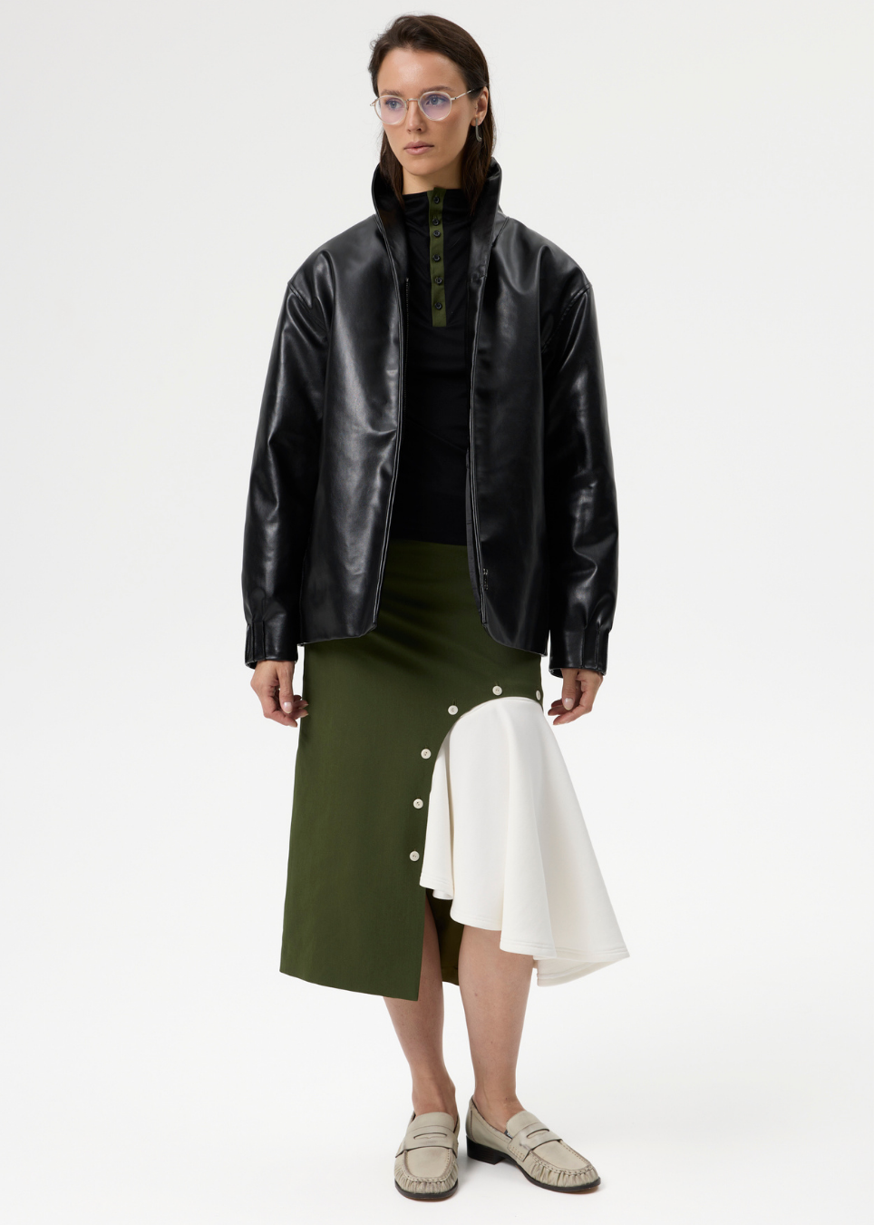 A model stands against a plain light gray studio background wearing a black leather-look jacket over a black top, paired with an olive green asymmetrical skirt featuring white side panels and buttons, and beige loafers.