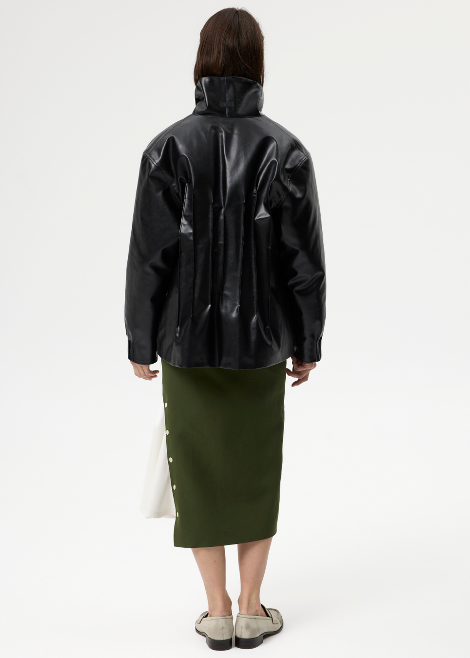 A model is shown from the back wearing a black glossy leather jacket, an olive green midi skirt, and beige loafers, holding a white bag against a plain white studio background.