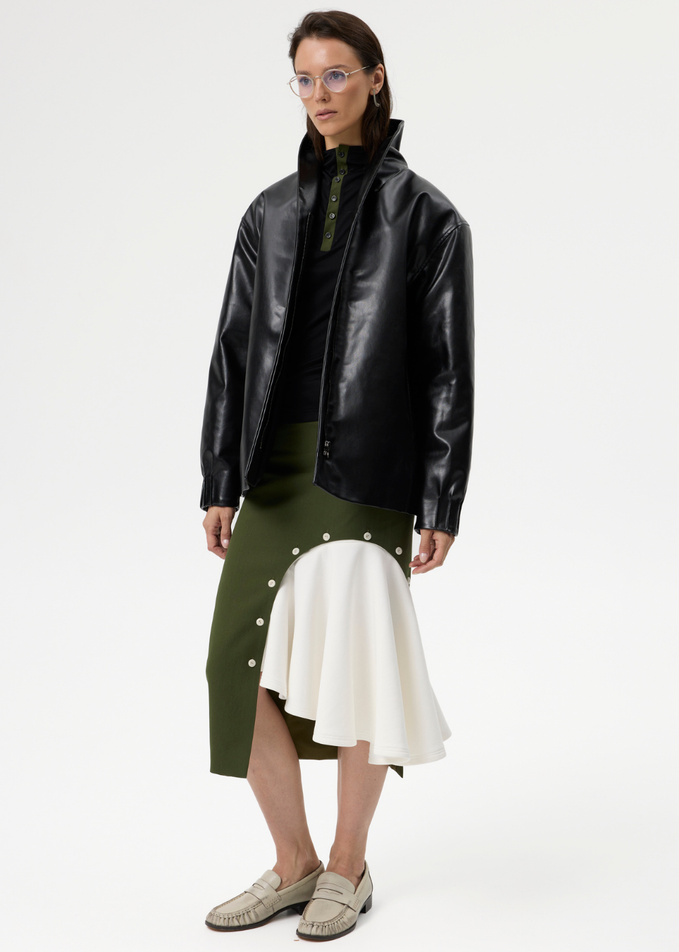 A model stands against a plain light gray studio background wearing a black leather jacket over a black top, paired with an asymmetrical olive green and white skirt and beige woven loafers.