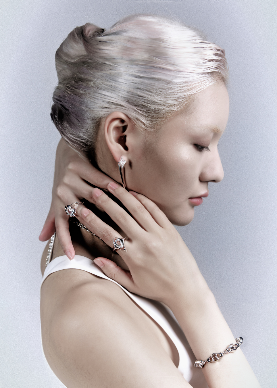 A model with platinum blonde hair styled in a bun, wearing delicate silver hoop earrings, rings, and a bracelet, posing with her hands near her neck against a light gray background.
