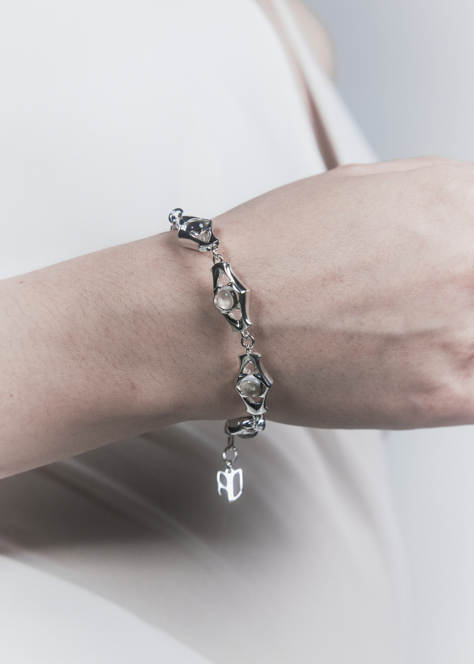 A close-up of a person's wrist wearing a silver-toned chain bracelet with irregular geometric links and a small dangling charm, against a soft light gray background.