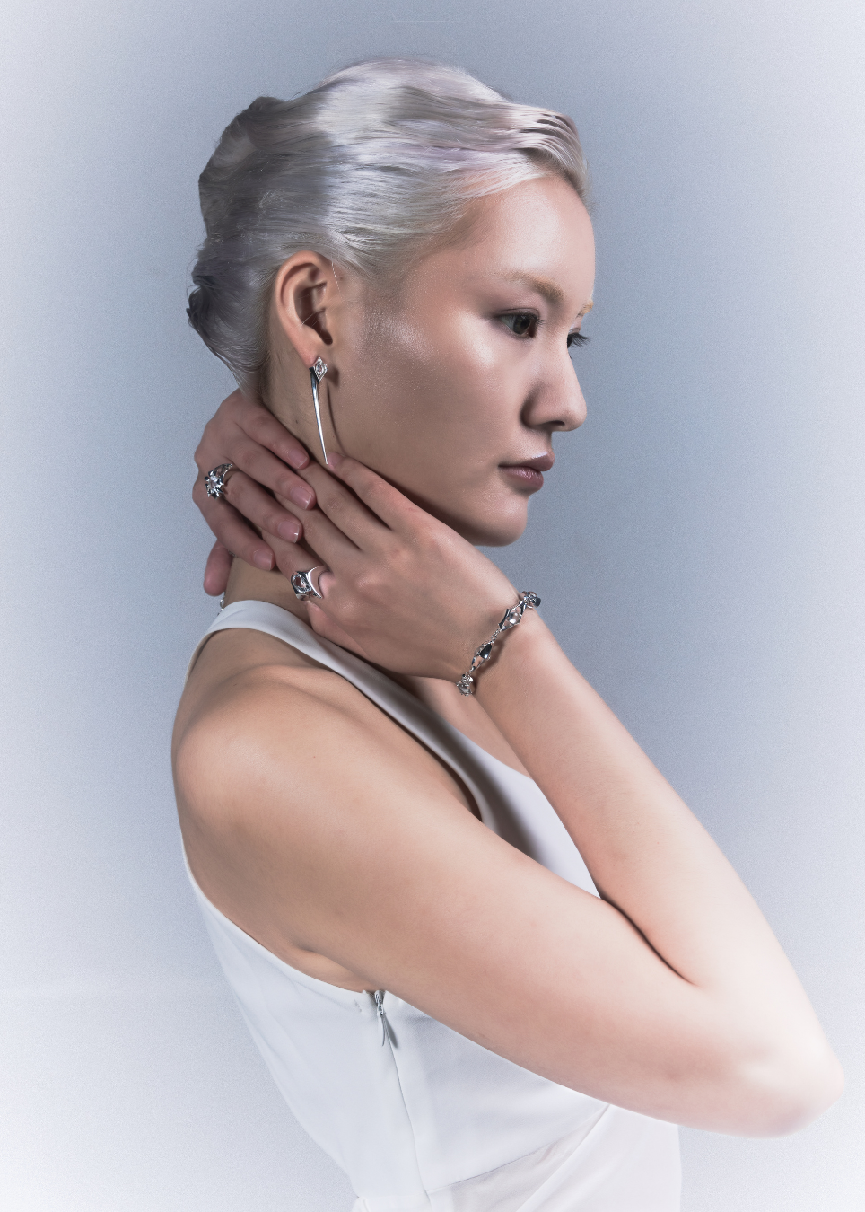 A model with short silver hair is shown in profile wearing a long silver hoop earring, a silver chain bracelet, and silver rings, posing with her hands near her neck against a light gray background.