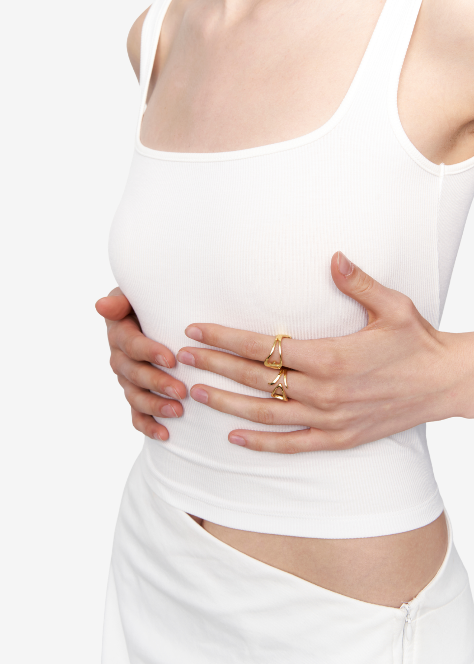 A close-up of a model wearing a white ribbed tank top and white bottoms, holding her stomach with her hands while wearing two gold rings.