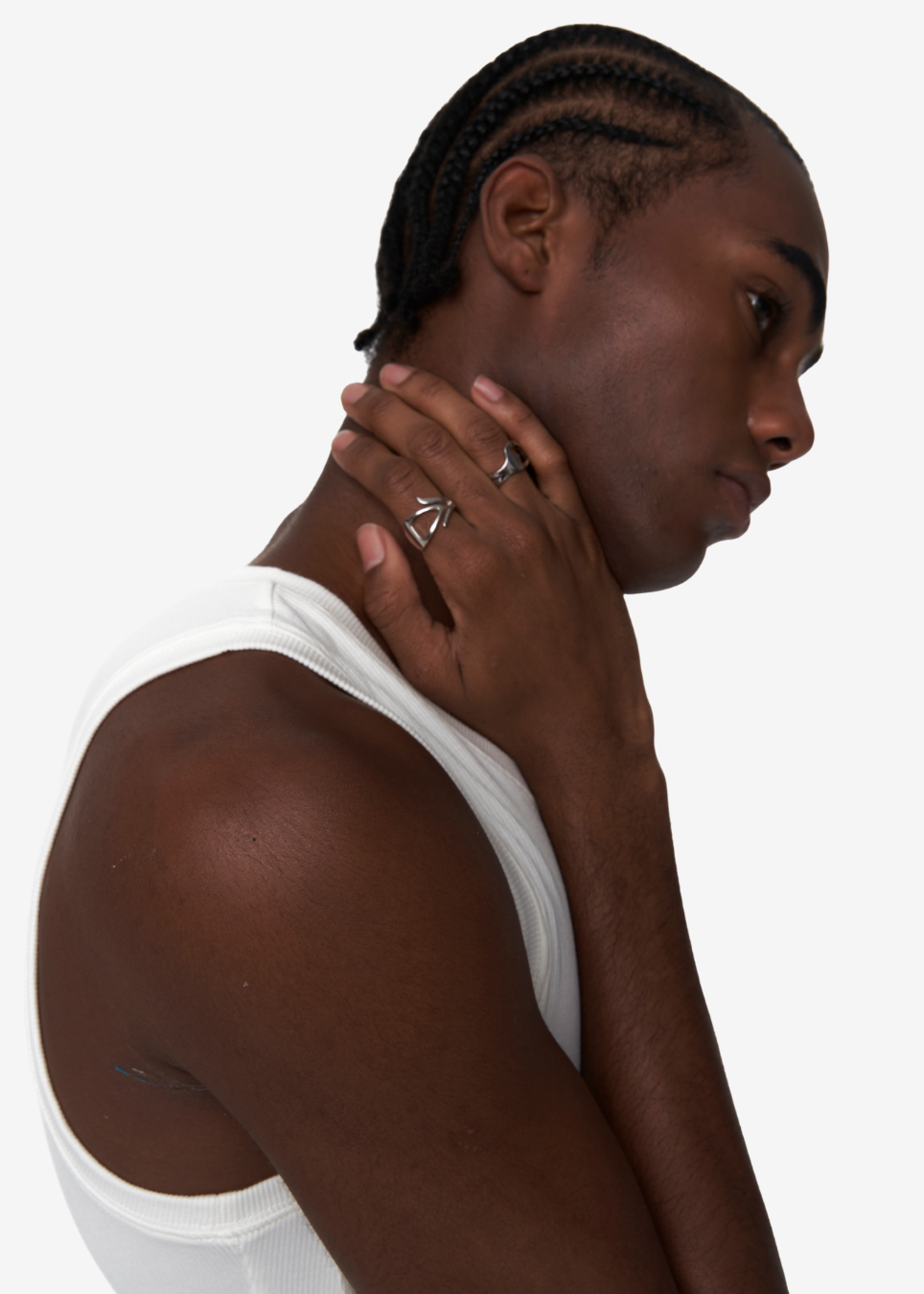 A man in profile wearing a white ribbed tank top and silver geometric rings on his fingers, set against a plain light gray background.