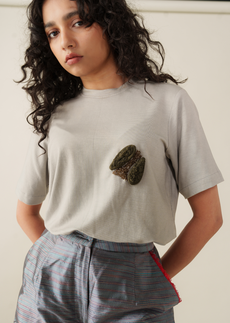 A model wearing a light gray t-shirt with a textured, organic-shaped green and brown patch on the chest, paired with striped gray shorts.