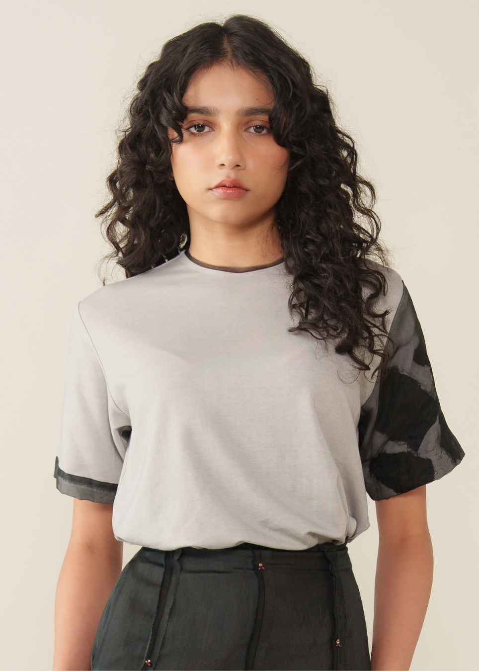 A model with long, curly dark hair wearing a light gray short-sleeve t-shirt with black patterned sheer sleeves and dark gray high-waisted pants, set against a plain beige background.