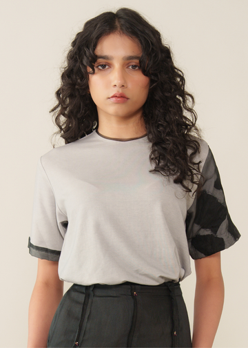 A model with long, curly dark hair wearing a light gray short-sleeve t-shirt with black patterned sheer sleeves and dark gray high-waisted pants, set against a plain beige background.