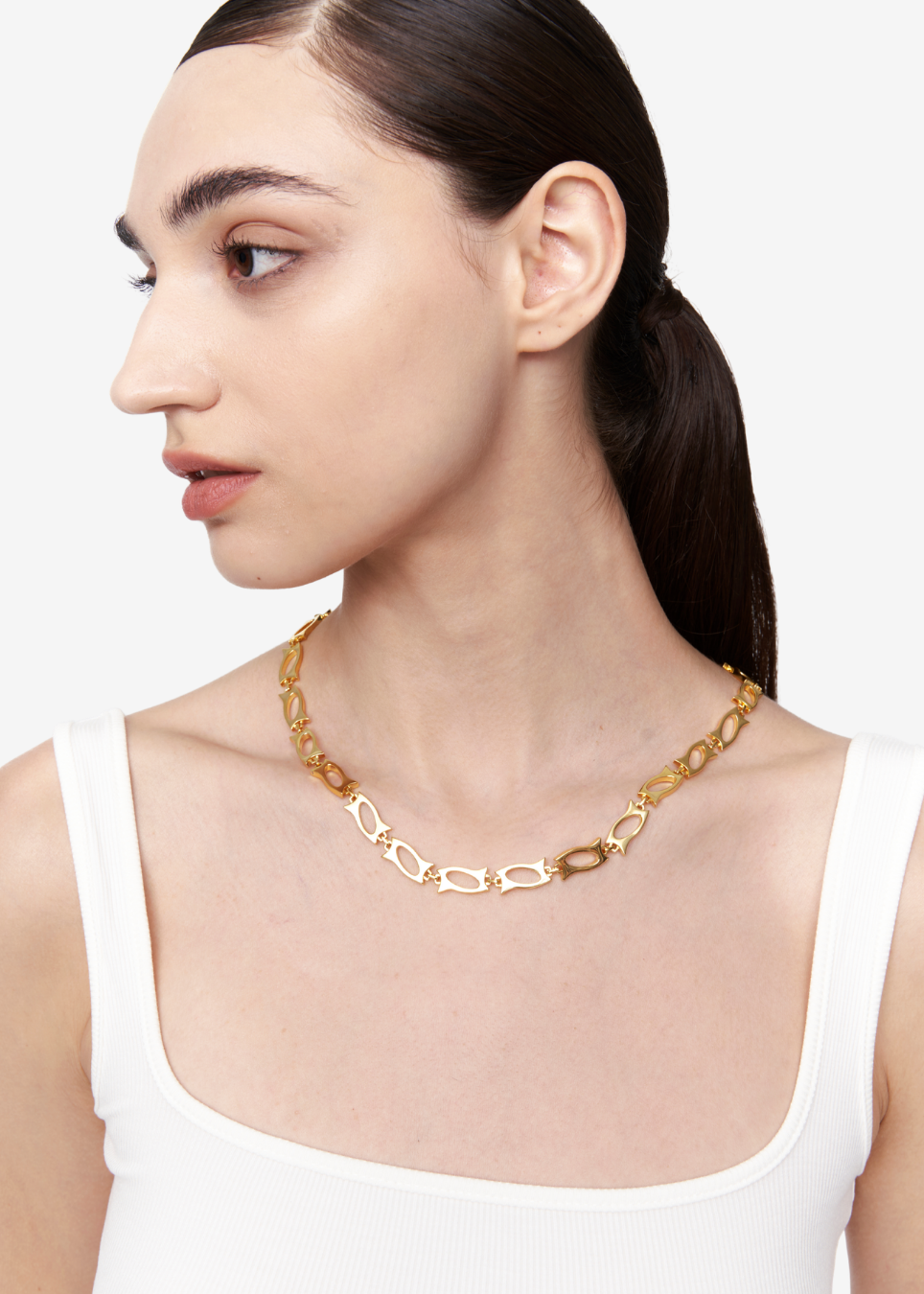 A close-up side profile of a woman wearing a gold chain necklace with elongated interlocking links, paired with a white ribbed tank top against a light gray background.