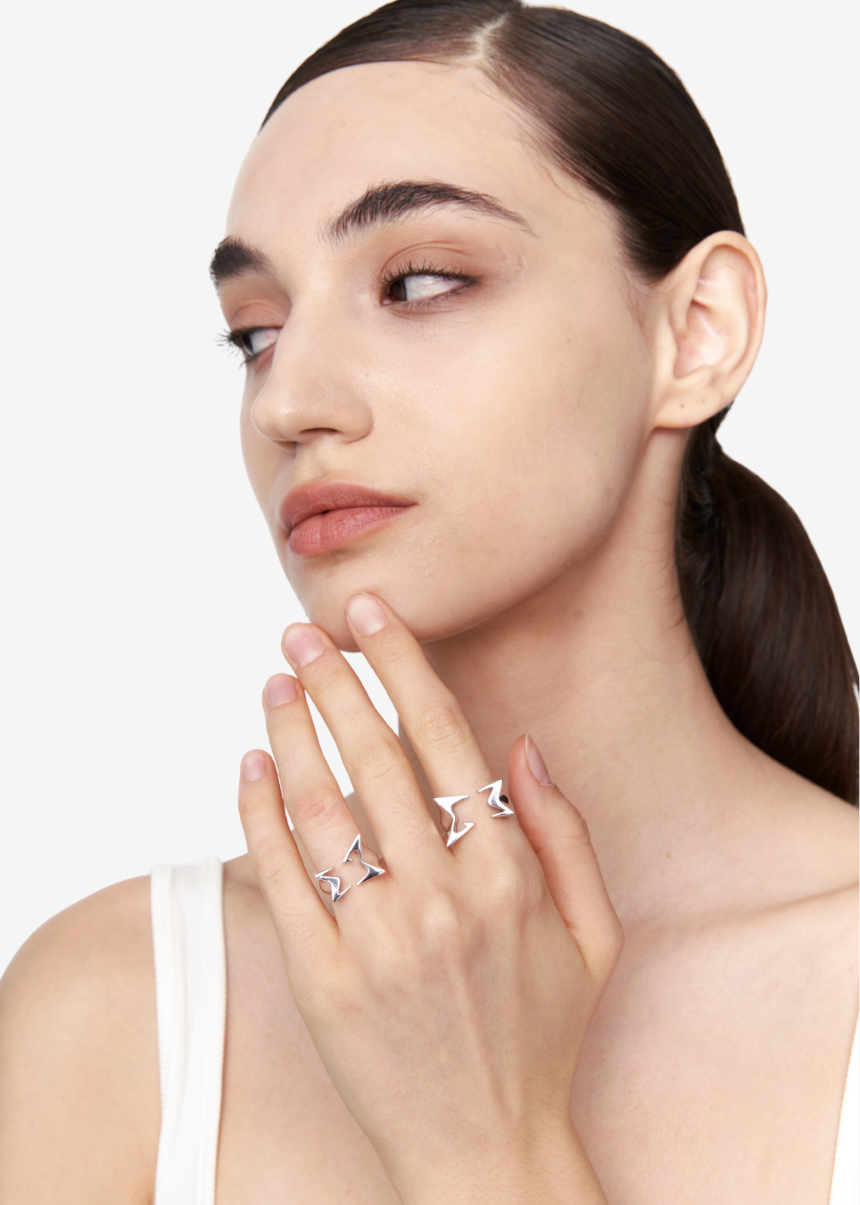 A close-up of a woman with her hair pulled back, wearing two silver geometric rings on her fingers, against a clean white background.